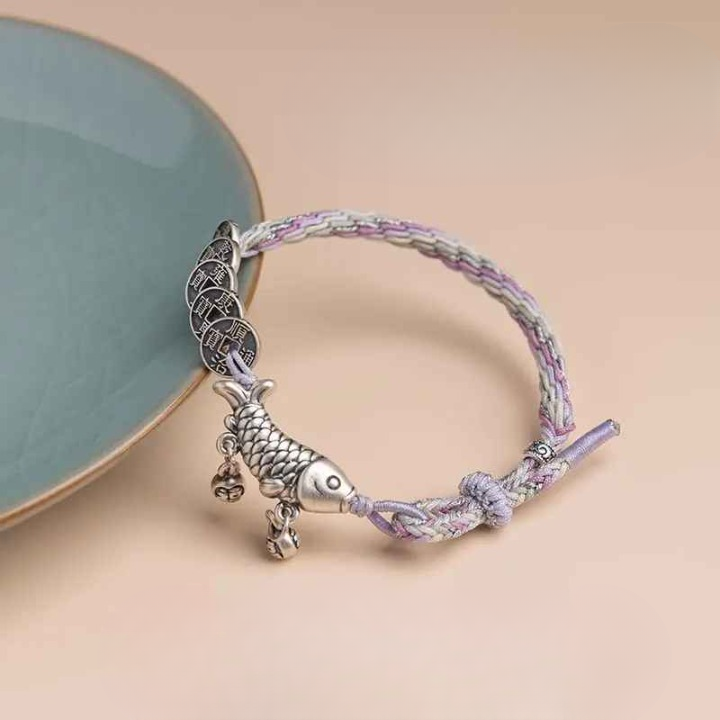 Bracelet carpe Koi et sapèques Feng Shui posé à plat sur fond beige, cordon tressé lilas et crème bien visible.